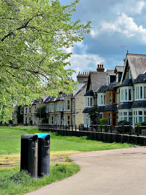 A row of Victorian-style terraced houses with bay windows and slate roofs, situated behind a well-maintained garden boundary fence. In the foreground, there is a paved pathway bordered by a lush, green lawn, and two black trash bins are placed near the path's edge. To the left, an overhanging tree with bright green leaves provides shade, its branches extending across the sky, which is partly cloudy with patches of blue visible. The scene appears to be in a suburban area where garden maintenance is evident, consistent with the services offered by Gardeners Merton, as seen in the context of Wimbledon Common gardeners serving Merton homes.